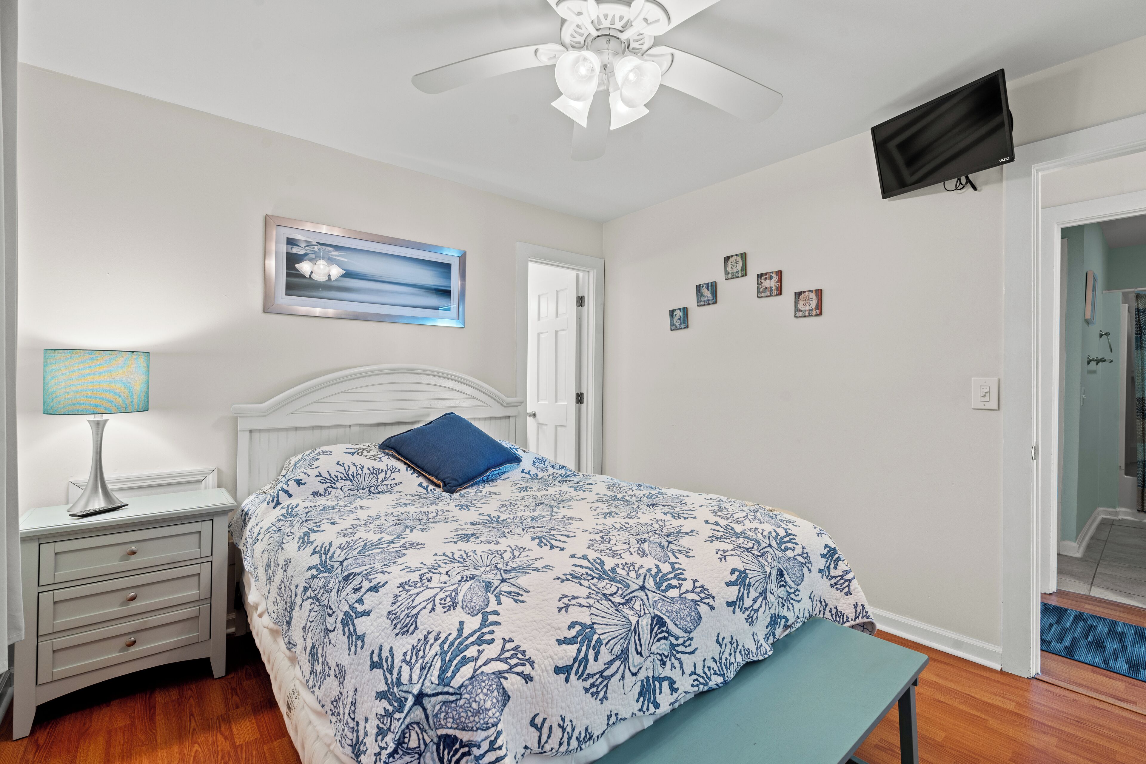 A bedroom with a bed, bedside table, lamp, ceiling fan, wall-mounted TV, and a door leading to another room.