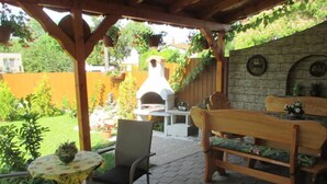 An outdoor patio with a stone floor, a wooden bench, a wicker chair, a small round table with a plant, and a pizza oven in the background. - Riviera Apartment Family Apartments near the swimming pool and aquapark! (Hajdúszoboszló)