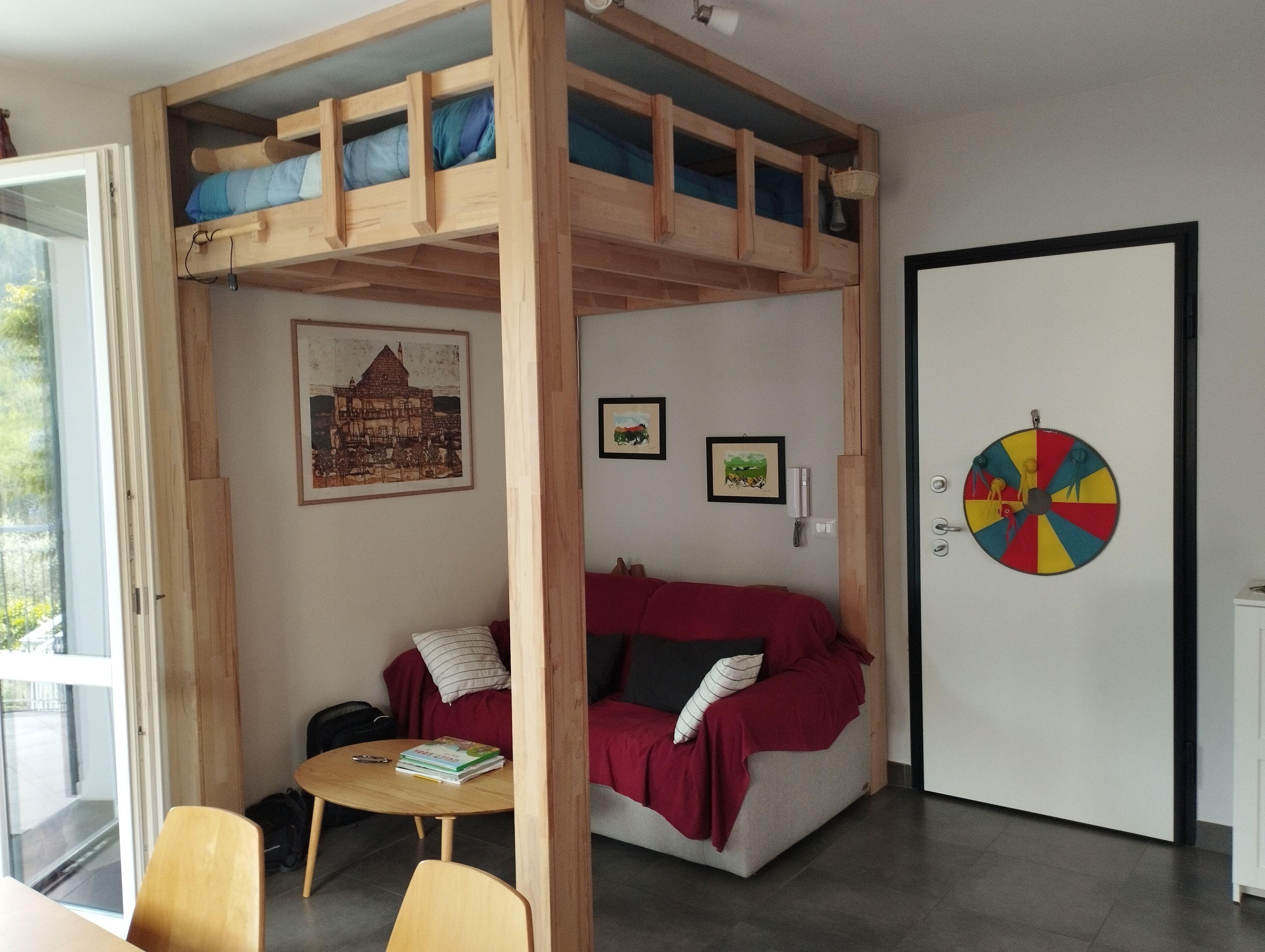 Une pièce avec un lit superposé, un canapé rouge, une petite table avec des livres et un jeu de fléchettes accroché au mur.