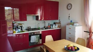 Une cuisine avec des placards rouges, une cuisinière et un évier. Il y a une horloge au mur et un panier de fruits sur la table.