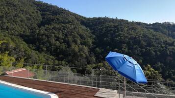 Une piscine avec une terrasse en bois, un parasol bleu et une vue sur une colline boisée.