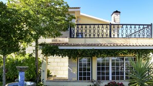 A two-story house with a balcony, a stone fountain, and a garden with various plants.