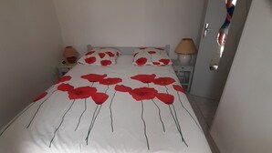 A bedroom with a bed covered in a white bedspread with red floral patterns, two bedside lamps, and a small wall decoration. - Coraux Residence house+Veranda park piscines (Canet-en-Roussillon)