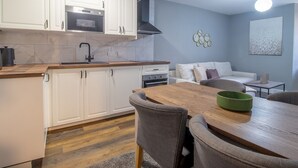 Cocina compacta con armarios blancos, horno integrado y estufa. Hay una zona de comedor con una mesa y sillas de madera, además de un sofá gris.