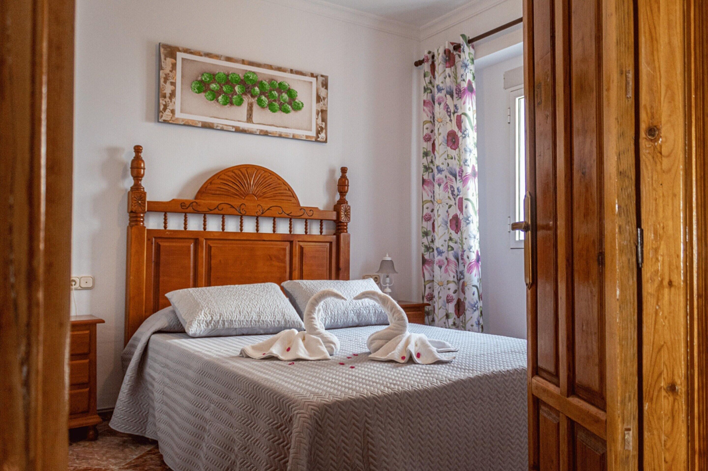 Un dormitorio con cabecera de madera, cama con colcha estampada, dos almohadas, mesita de noche y un cuadro enmarcado en la pared, junto con una cortina con motivos florales.