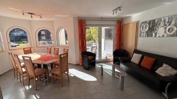 A modern living room with a dining area, large windows, and a sliding glass door leading to an outdoor patio.