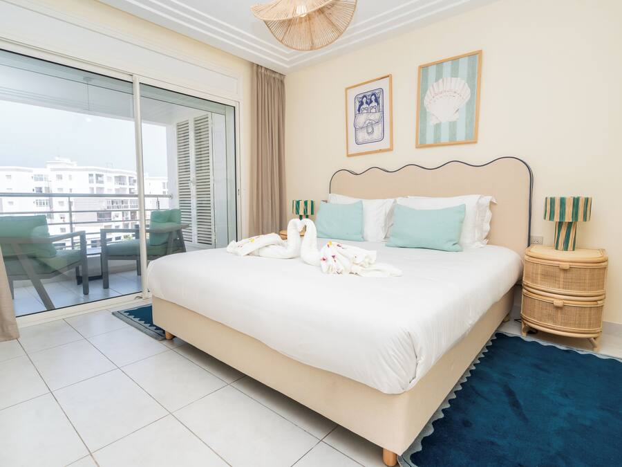 Habitación de hotel con una cama grande, un balcón con vistas a edificios y una mesita de noche de mimbre con lámpara.