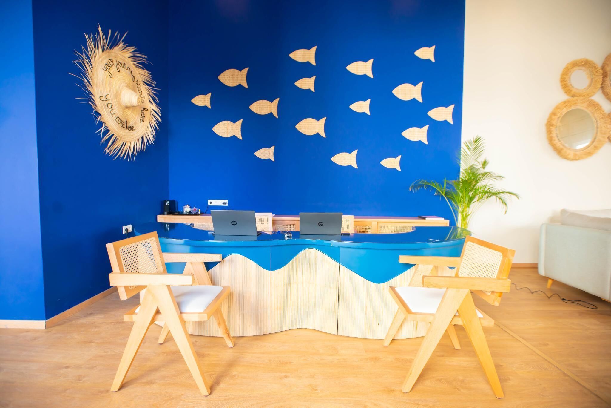 A reception desk with two HP laptops, wooden chairs, a blue wall with fish cutouts, and a straw hat decoration.