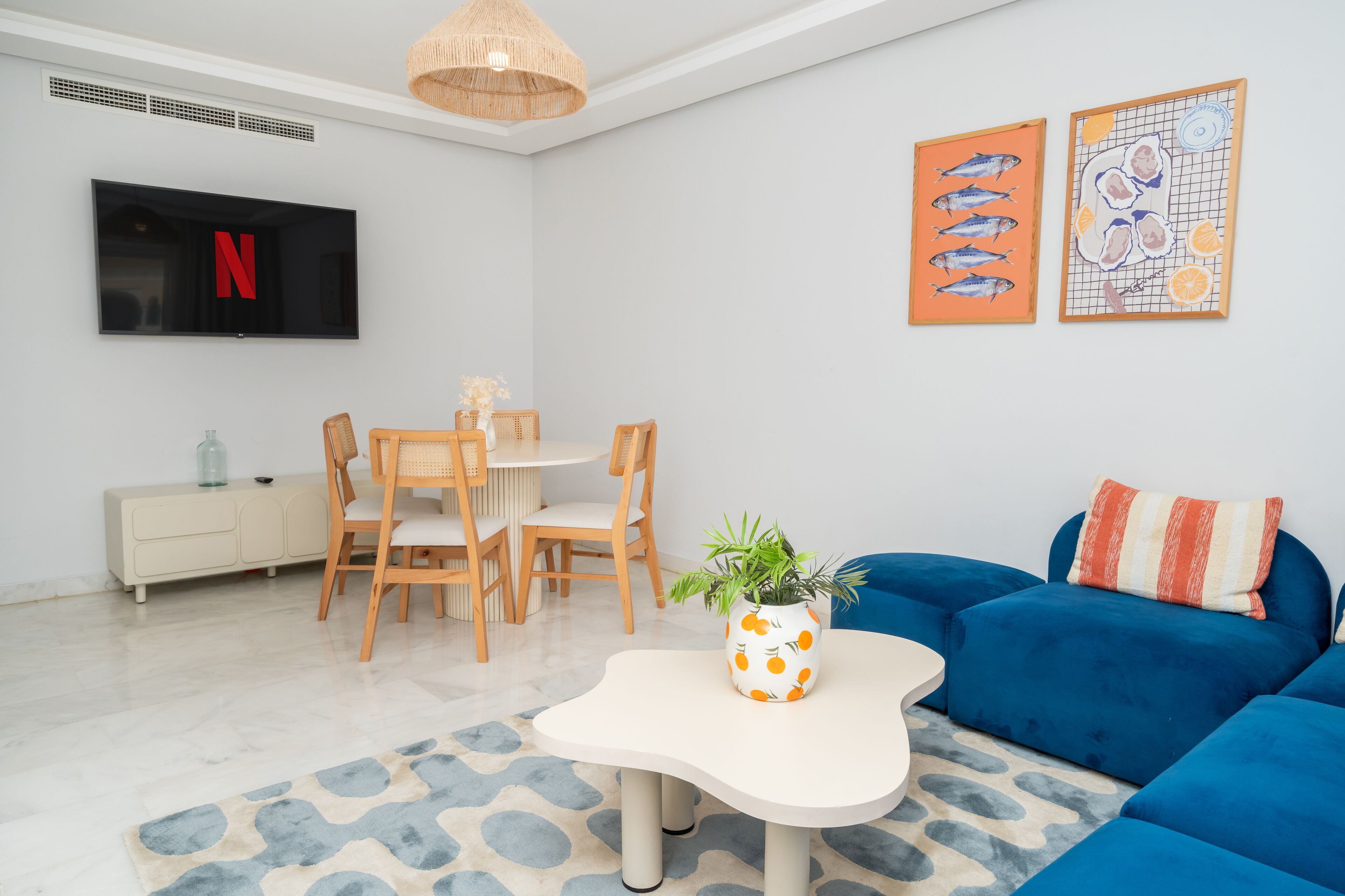 Una sala de estar moderna con televisor, mesa de comedor, sillas, un sofá azul y obras de arte en las paredes.