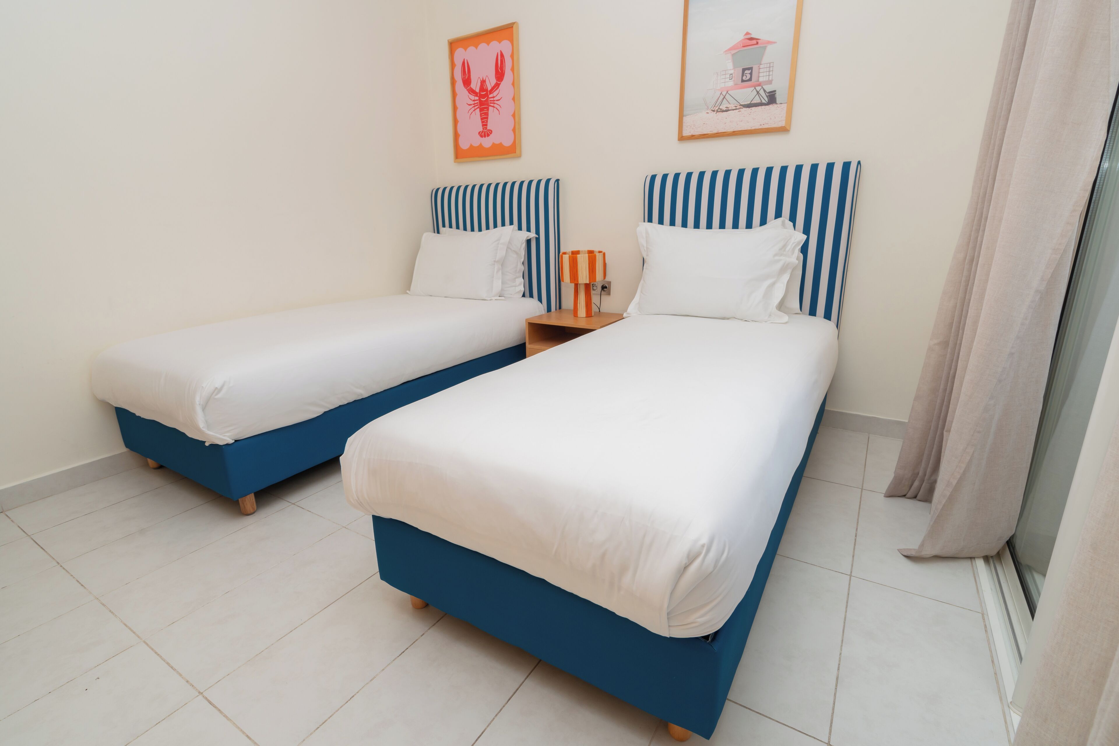 A hotel room with two beds, each with a blue headboard and white bedding. There are two framed pictures on the wall, one of a lobster and another of a beach scene.