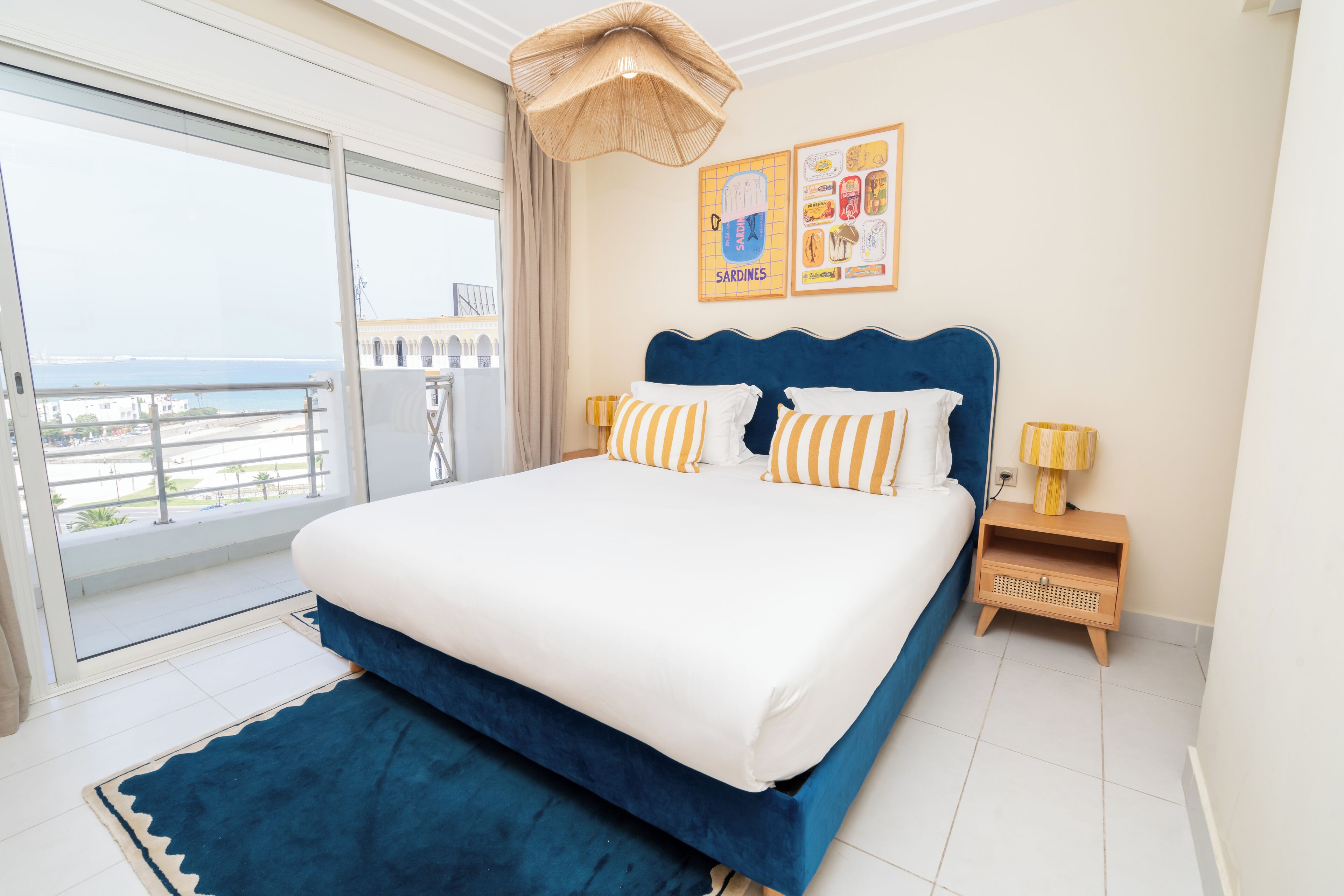 A bedroom with a large bed, a view to the outside, a small table with a lamp, and a wall with framed pictures.