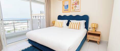 A bedroom with a large bed, a view to the outside, a small table with a lamp, and a wall with framed pictures.