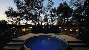 Ein Schwimmbad mit Whirlpool, umgeben von Stufen und Gartenmöbeln, mit Bäumen und einem beleuchteten Zaun im Hintergrund.