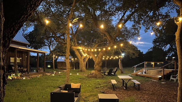 Ein Garten mit einer Terrasse, Lichterketten, Holzmöbeln und einer überdachten Sitzgelegenheit.