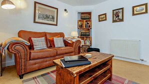 A cozy living room with a brown leather sofa, a wooden coffee table, and a bookshelf filled with books. - Jacobite's Retreat, 17th century cottage (Inverness)