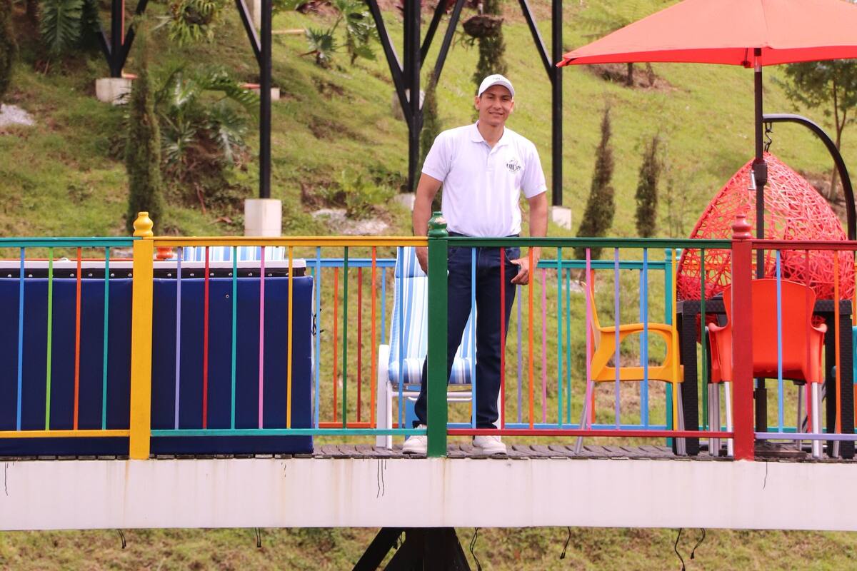 A man standing on a balcony with a colorful railing, overlooking a garden with various plants and a building in the background.