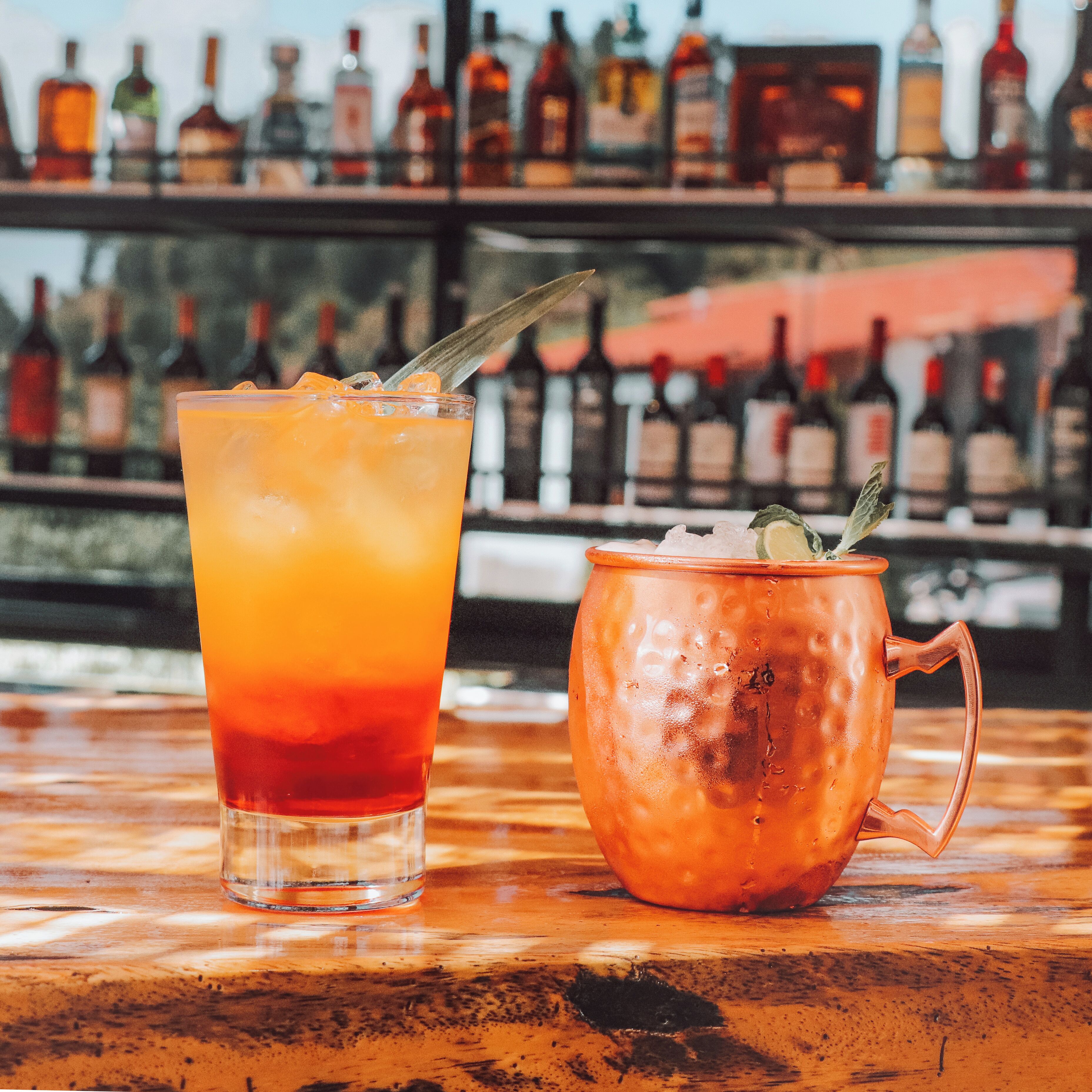 Two drinks on a bar counter with a background of bottles.