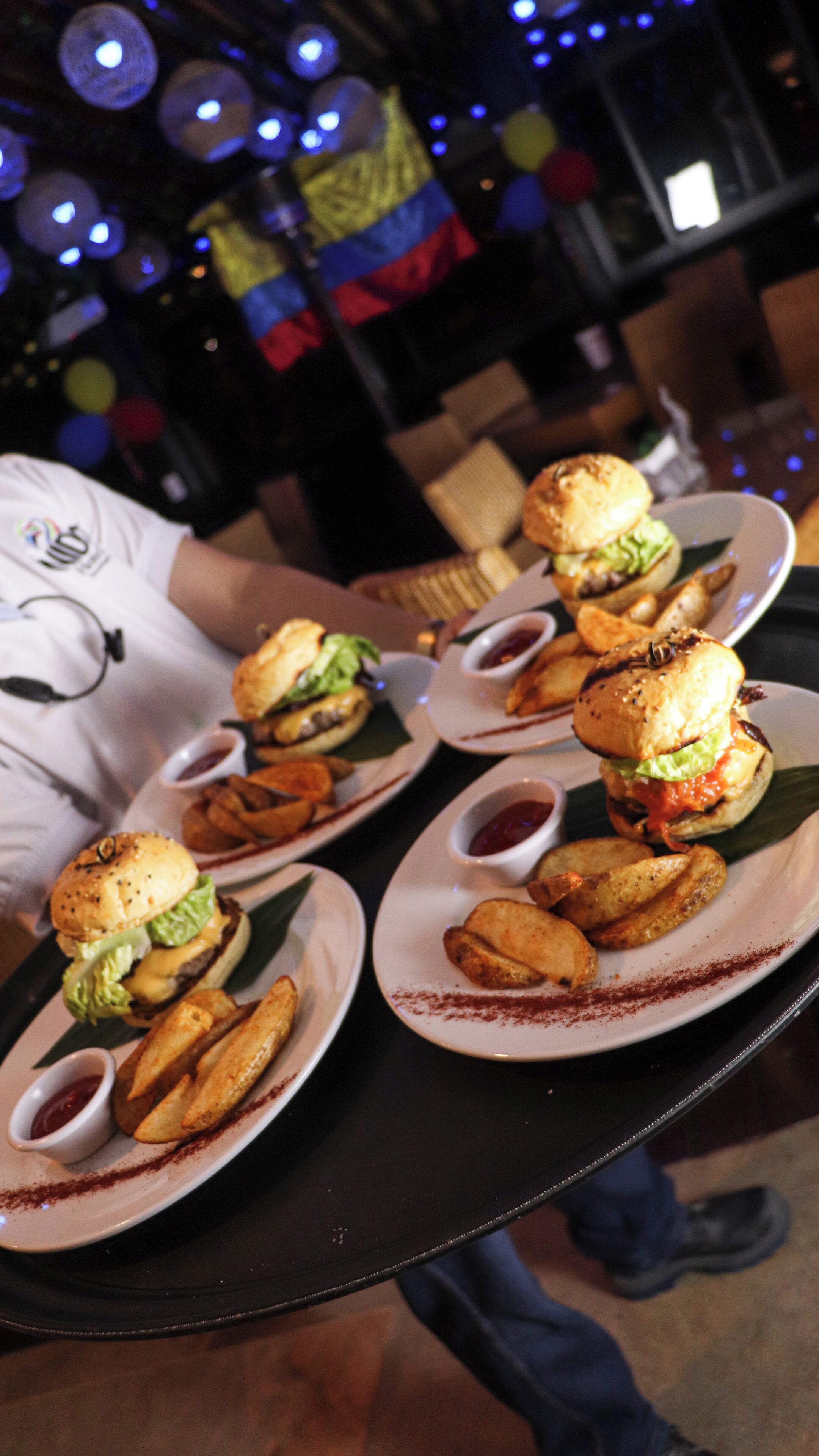 A table with plates of food, including burgers and fries, and a person in a white shirt holding a plate.