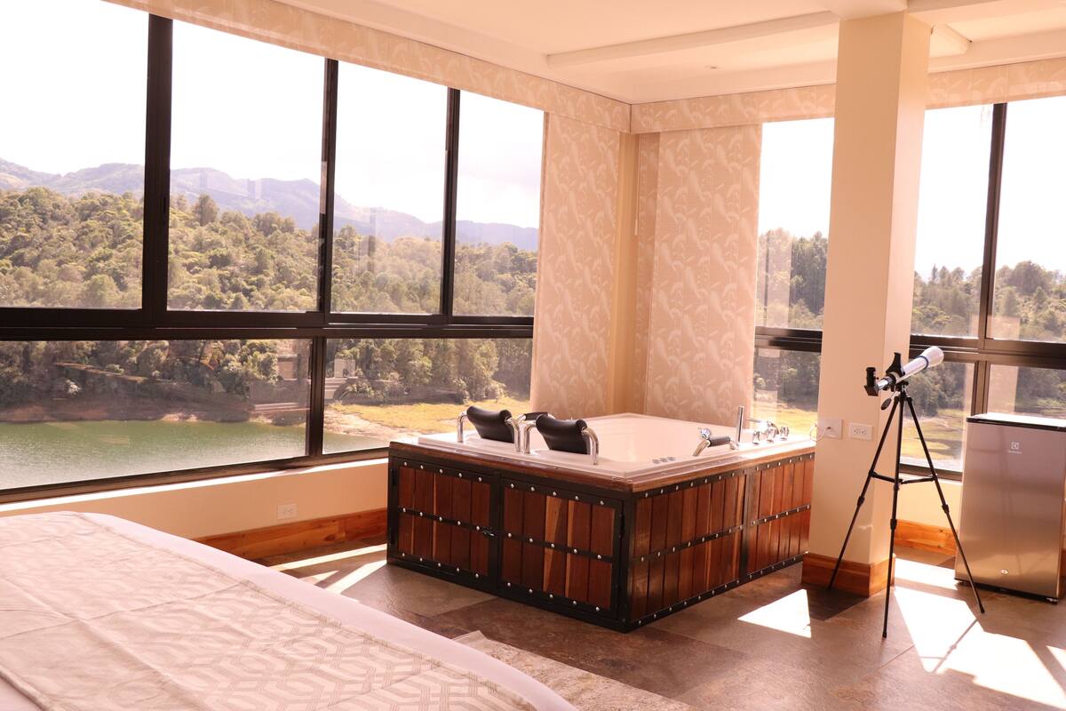 A bedroom with a large window, a wooden bathtub, and a tripod-mounted telescope.