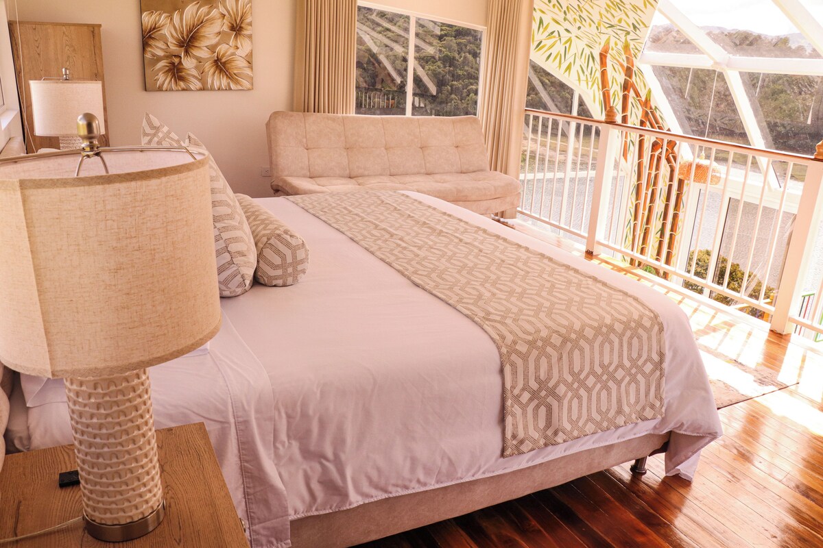 A bedroom with a large bed, a bedside table, a lamp, a sofa, and a view of a balcony with bamboo plants.