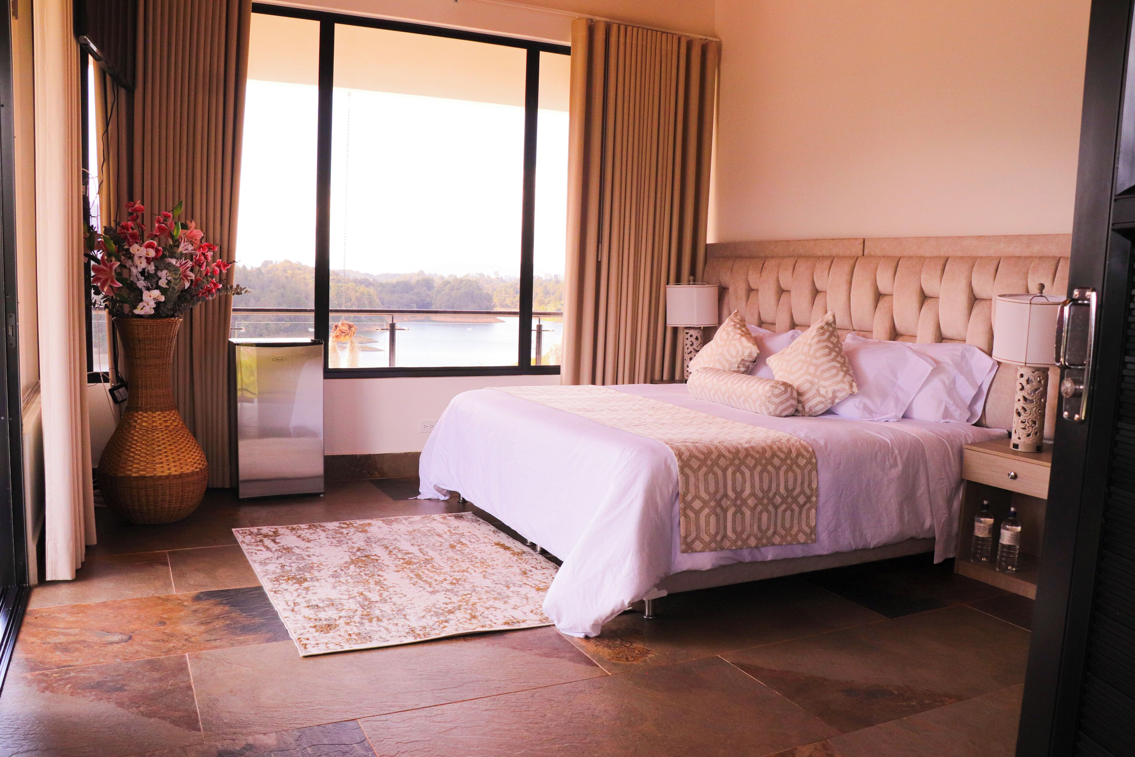 A bedroom with a large bed, a view of a lake, a vase of flowers, and a nightstand with a lamp.