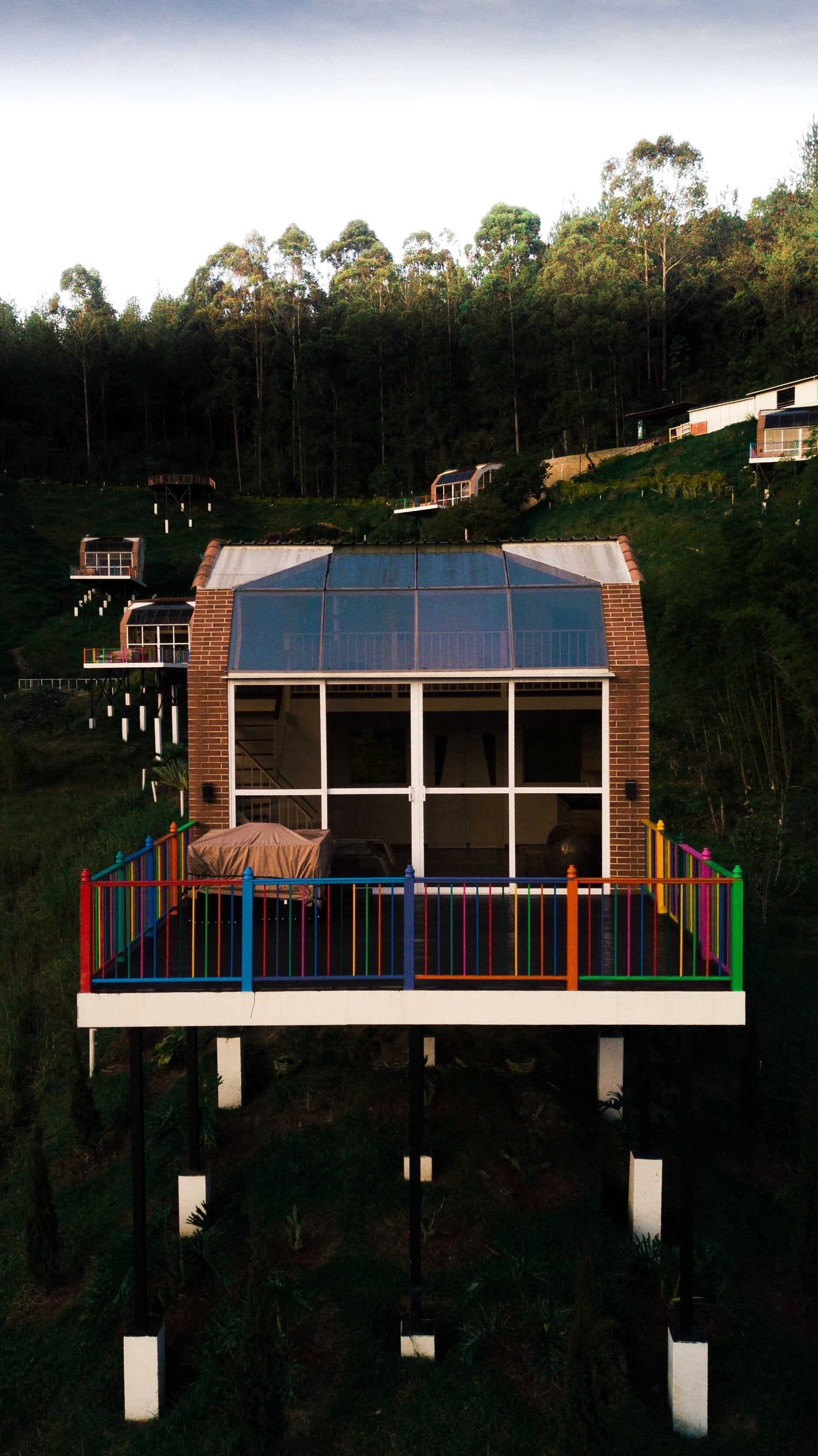 A modern house with a glass facade, elevated on stilts, surrounded by a colorful fence, and set in a forested area.