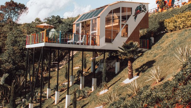 Una casa moderna con facciata in vetro, sollevata su palafitte, immersa nel verde e con cielo sereno.