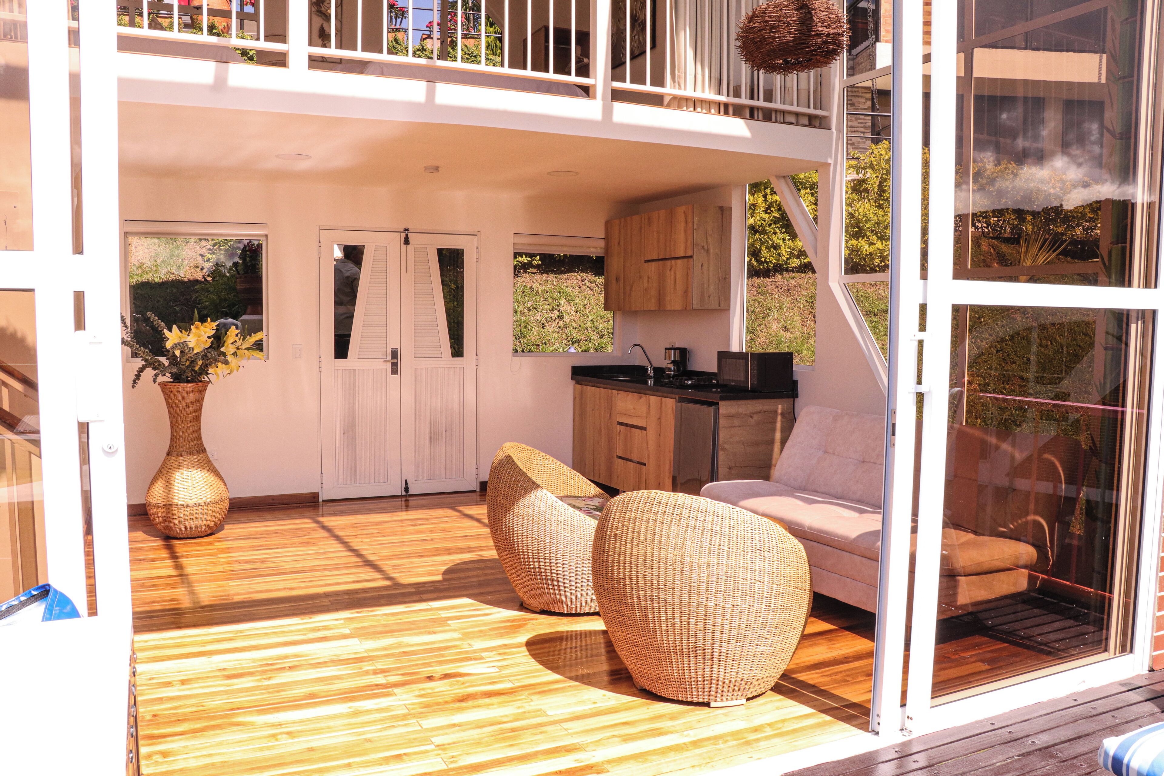 A modern interior with a wooden floor, wicker furniture, a kitchen area, and large windows.