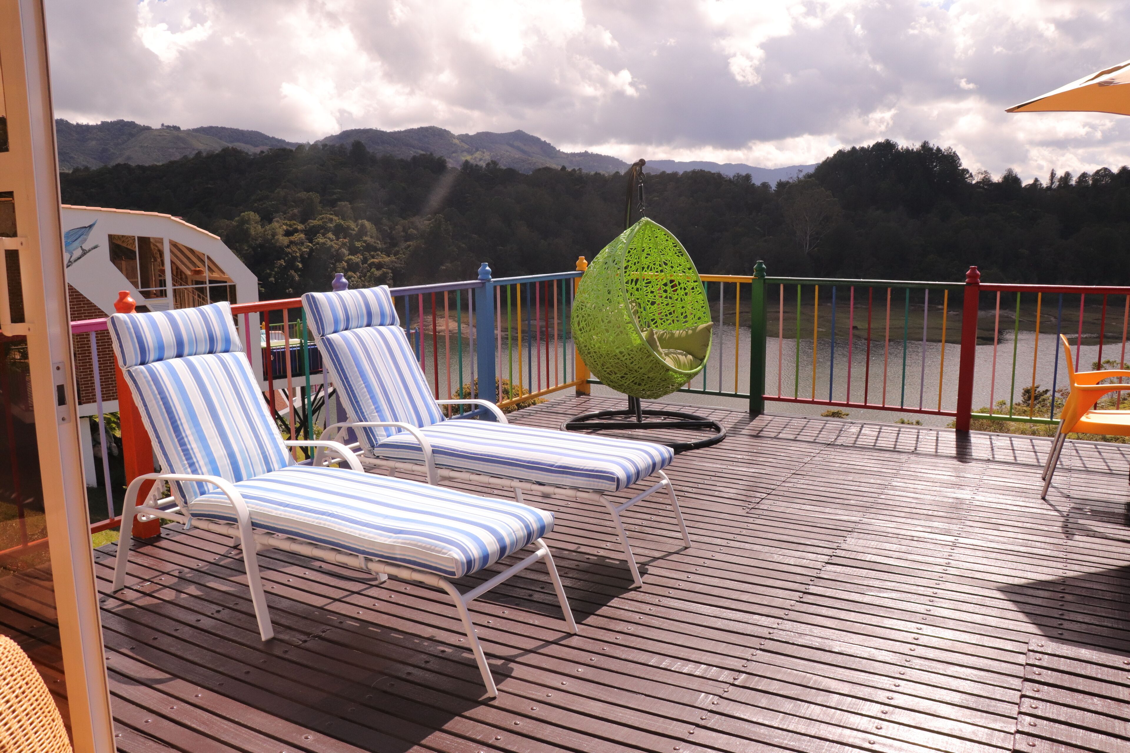 A deck with striped lounge chairs and a hanging chair overlooking a body of water and forested hills.