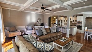 Een ruime woonkamer met een bruine leren bank, decoratieve kussens en een houten salontafel. Een eethoek met witte stoelen en een houten tafel is zichtbaar. De keuken heeft witte kasten en een groot eiland.