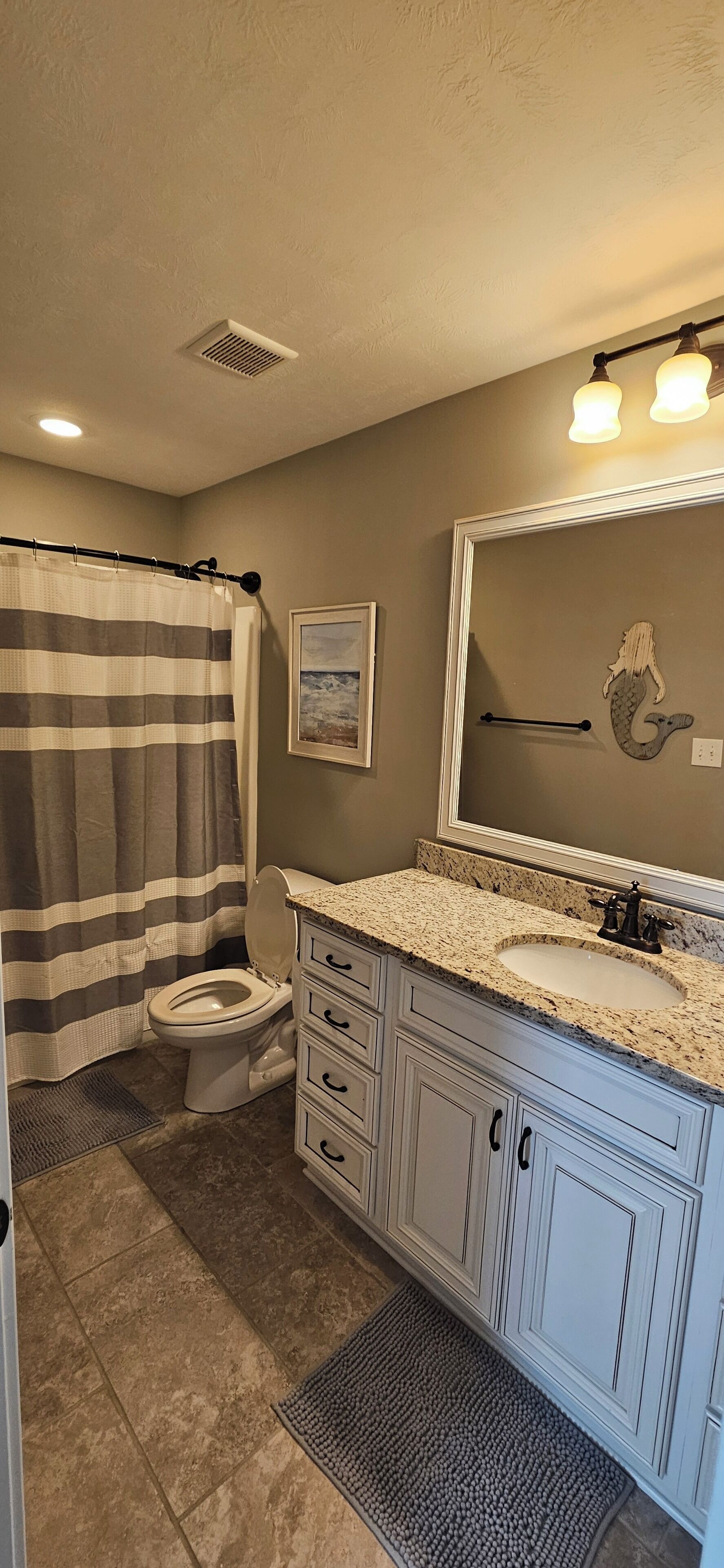 A bathroom with a striped shower curtain, a toilet, a sink with a granite countertop, and a mirror with a decorative frame.