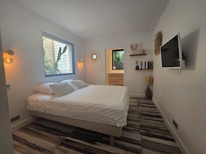 Een slaapkamer met een bed, een raam, een televisie en een zichtbare badkamer op de achtergrond.