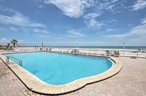 A rectangular swimming pool with a brick border, surrounded by a paved deck, and overlooking a beach with a clear sky.