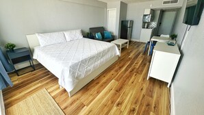 A bedroom with a large bed, a desk, a chair, a small table, a mirror, and a kitchenette.