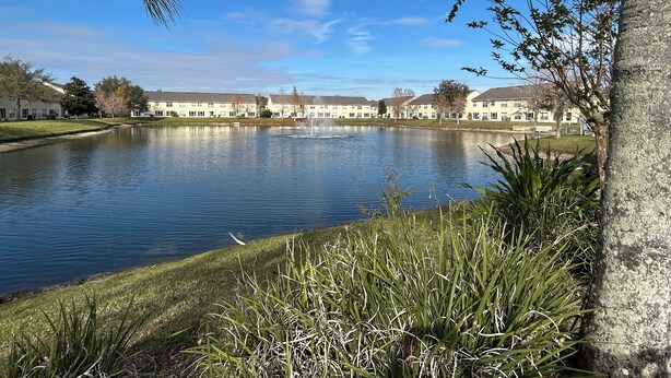A tranquil lake with a fountain, surrounded by a well-maintained lawn and a row of buildings with a uniform architectural style. - ★12Mins to Disney Theme Parks- Save 25pct Off Stay- Pool- 3BR Condo-Sleeps 10★ (Kissimmee)