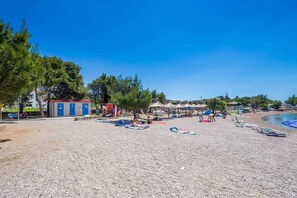 A beach with a row of blue cabins, a sandy area with umbrellas, and a clear blue sky.