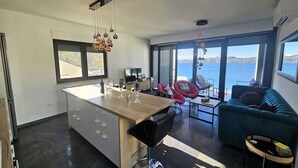 Eine moderne Küche mit einer zentralen Kochinsel, einem Essbereich mit Stühlen, einer Couch und Blick auf das Meer.