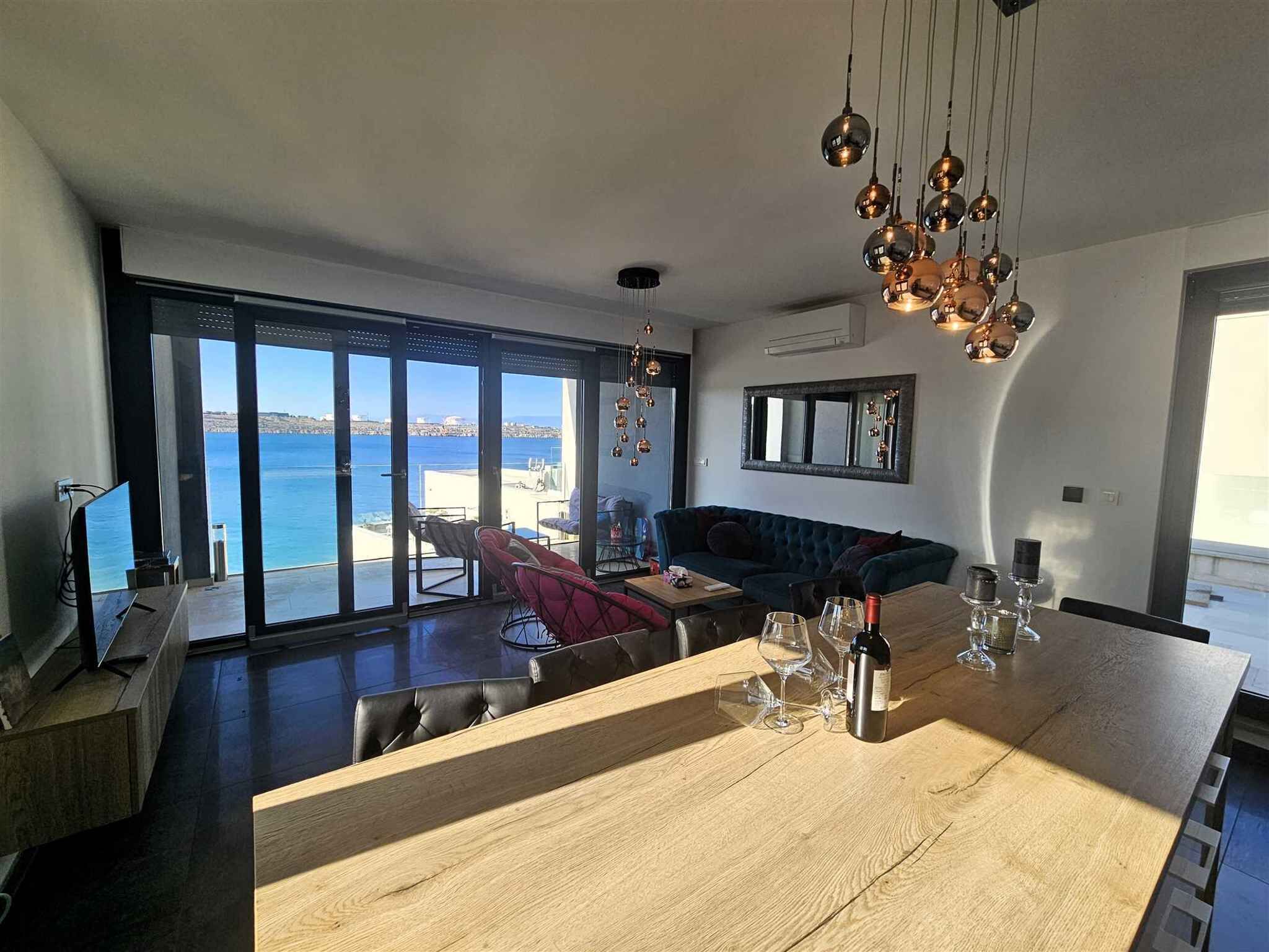 Een moderne woonkamer met een grote houten eettafel, een bank en uitzicht op zee door grote ramen.