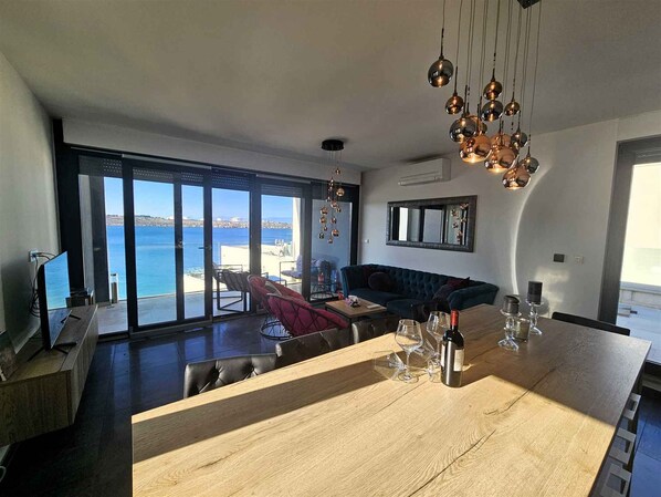 A modern living room with a large wooden dining table, a sofa, and a view of the sea through large windows.