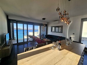 Un salon moderne dotĂ© dâune grande table Ă manger en bois, dâun canapĂ© et offrant une vue sur la mer grĂące Ă de grandes fenĂȘtres.