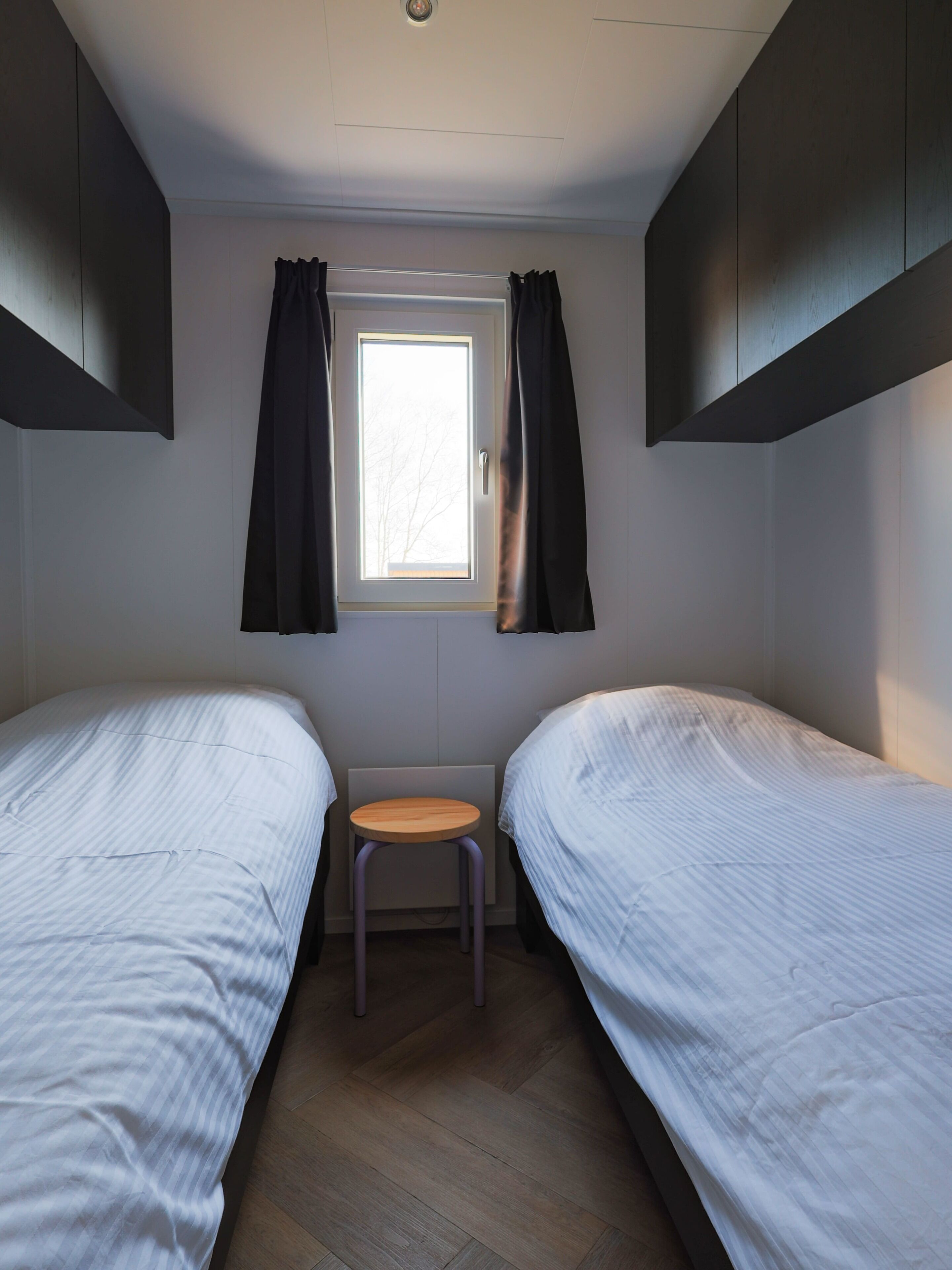 Een kleine, moderne kamer met twee eenpersoonsbedden, een tafeltje en een raam met gordijnen.
