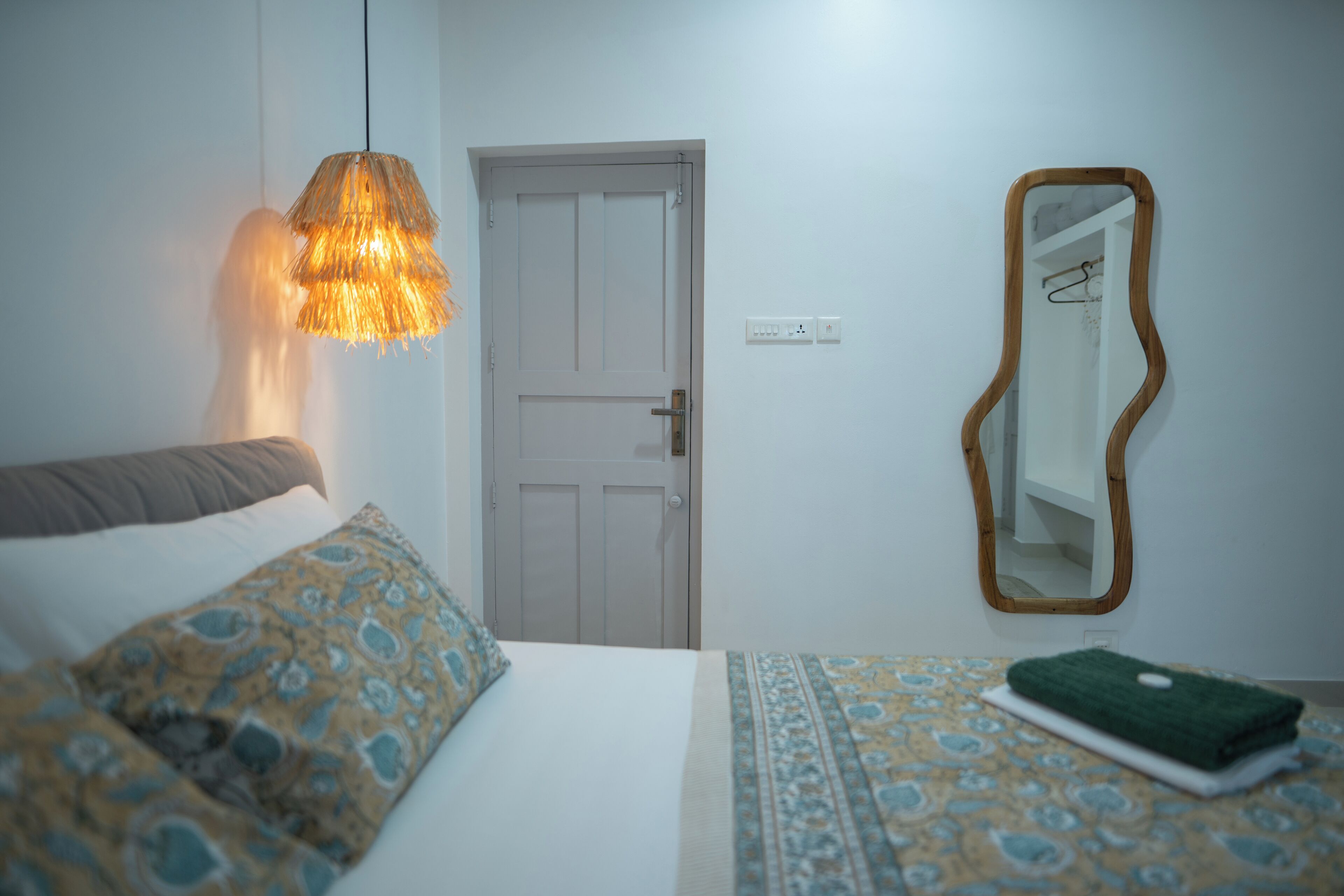 A bedroom with a bed, patterned pillows, a hanging light fixture, a door, and a mirror on the wall.