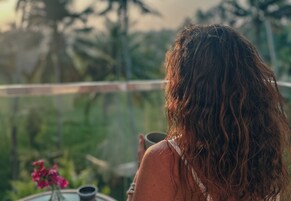 Una persona de cabello largo sostiene una taza, mirando un balcón con vistas a la vegetación y una mesa con un jarrón de flores.