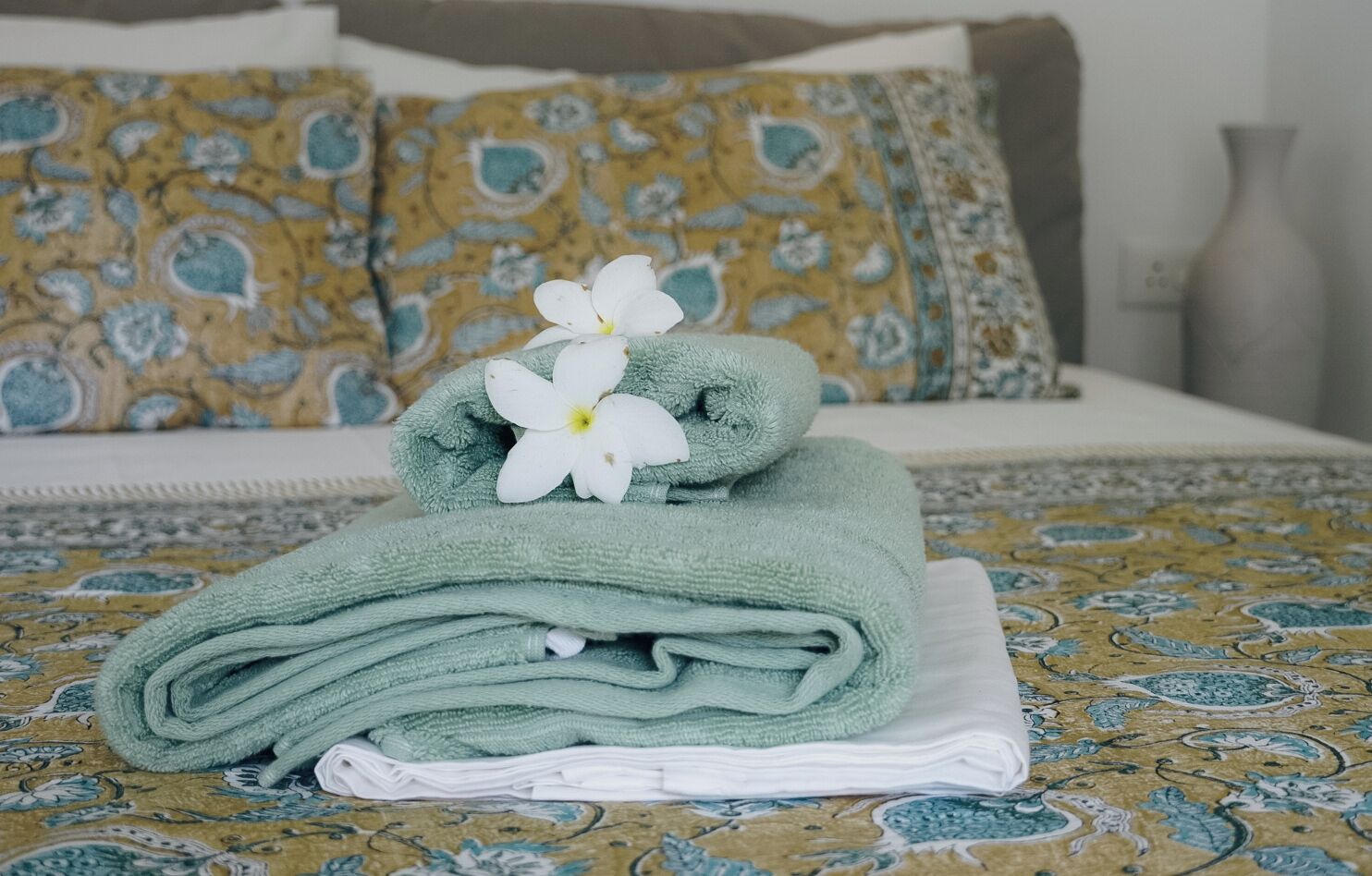 A neatly folded green towel with a white flower on a bed with a patterned bedspread.