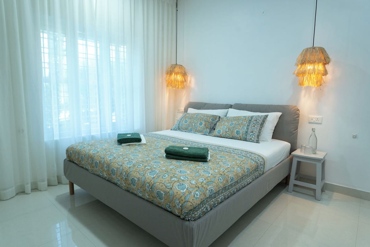 A modern bedroom with a large bed, two bedside lamps, a nightstand, and sheer curtains.