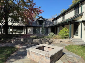 A courtyard with a stone fire pit, a stone retaining wall, and a house with a gabled roof. - Glen Flora; Geneva Lake Guest House with Private Pier, Fire Pit, Pool Table (Williams Bay)
