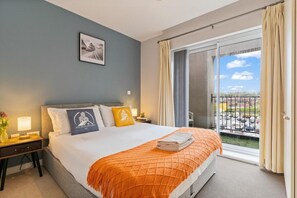 Habitación de hotel con cama, mesita de noche, lámpara, un cuadro en la pared y un balcón con vista al estacionamiento.