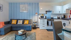 Una sala moderna con sofá, mesa de centro y una mesita auxiliar. Se ve una cocina con gabinetes y electrodomésticos blancos al fondo.