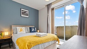 Un dormitorio con cama, mesita de noche, lámpara, un cuadro en la pared y ventana con cortinas.