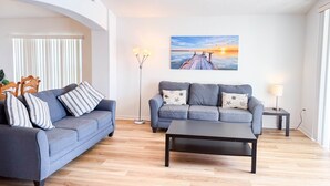 A living room with two sofas, a coffee table, a floor lamp, a ceiling fan, and a sunset painting on the wall.